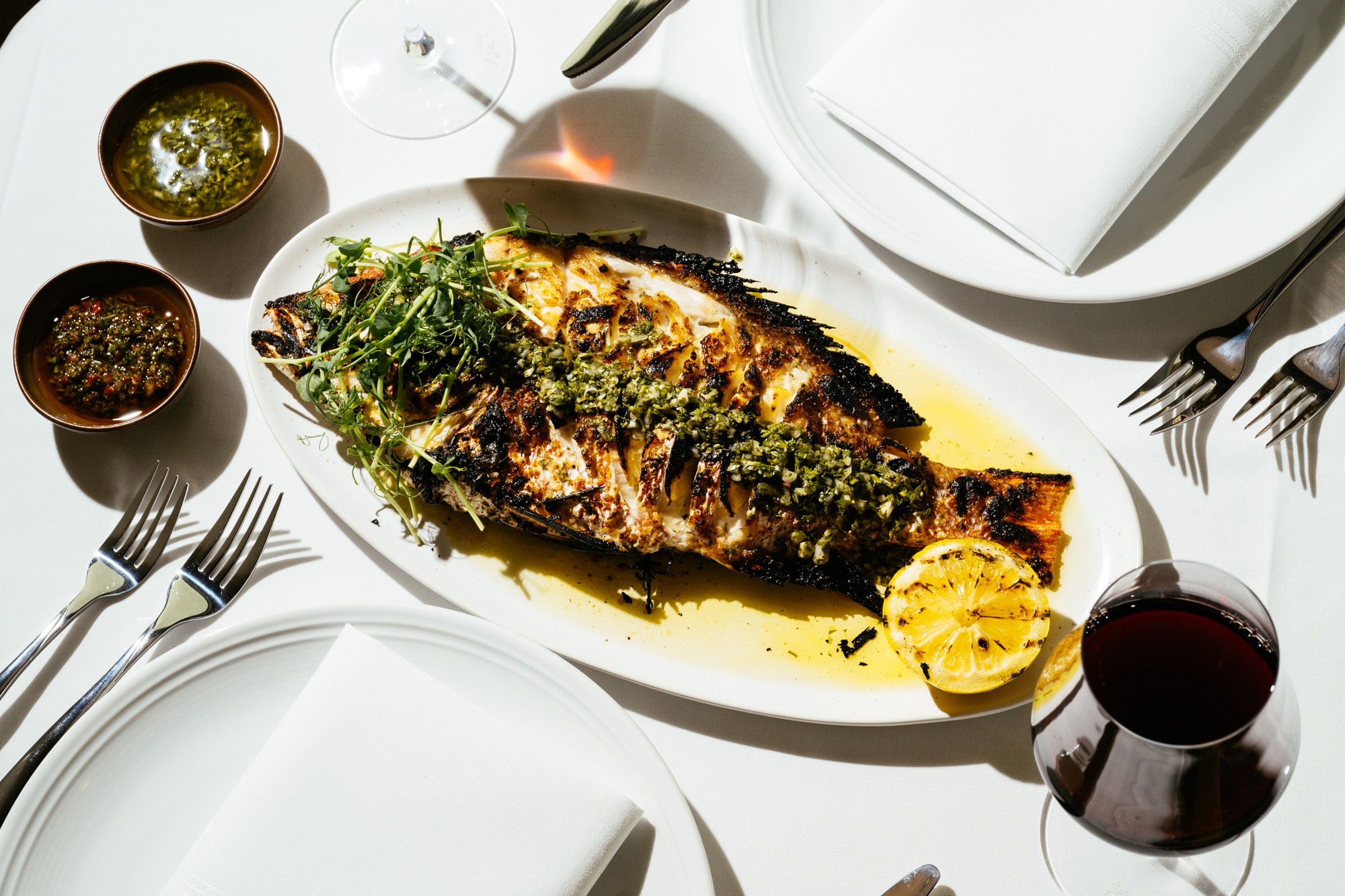 A whole grilled fish with herbs and charred lemon on a white tablecloth at Brasa, surrounded by sauces, silverware, and a glass of red wine