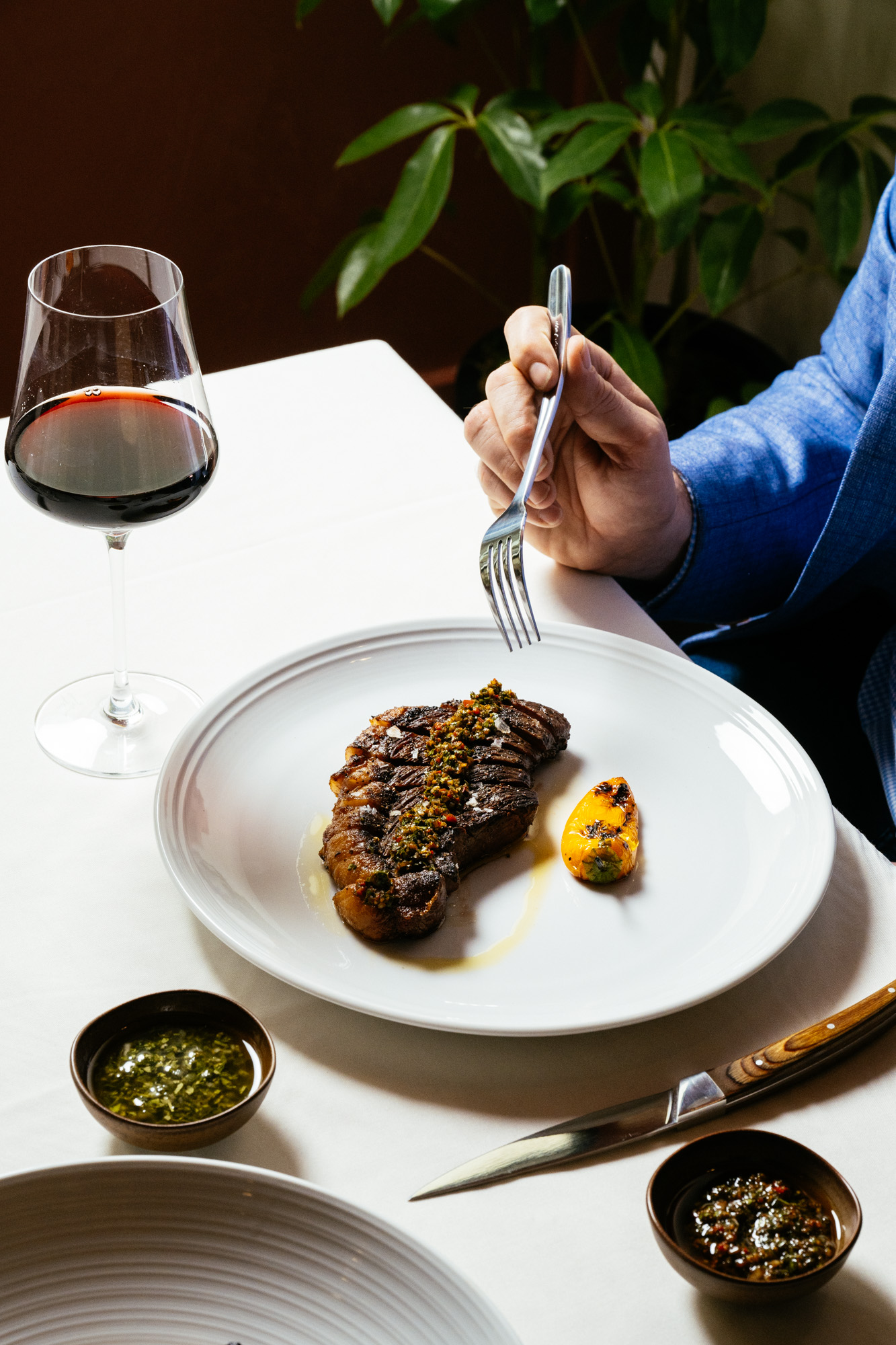 A guest in a blue jacket dining on steak with a glass of red wine at a white-clothed Brasa table
