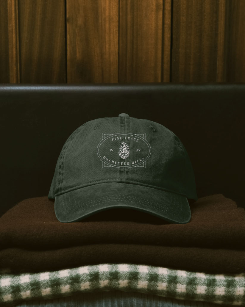 A dark green Pine Trace cap embroidered with the pinecone crest, resting on folded garments